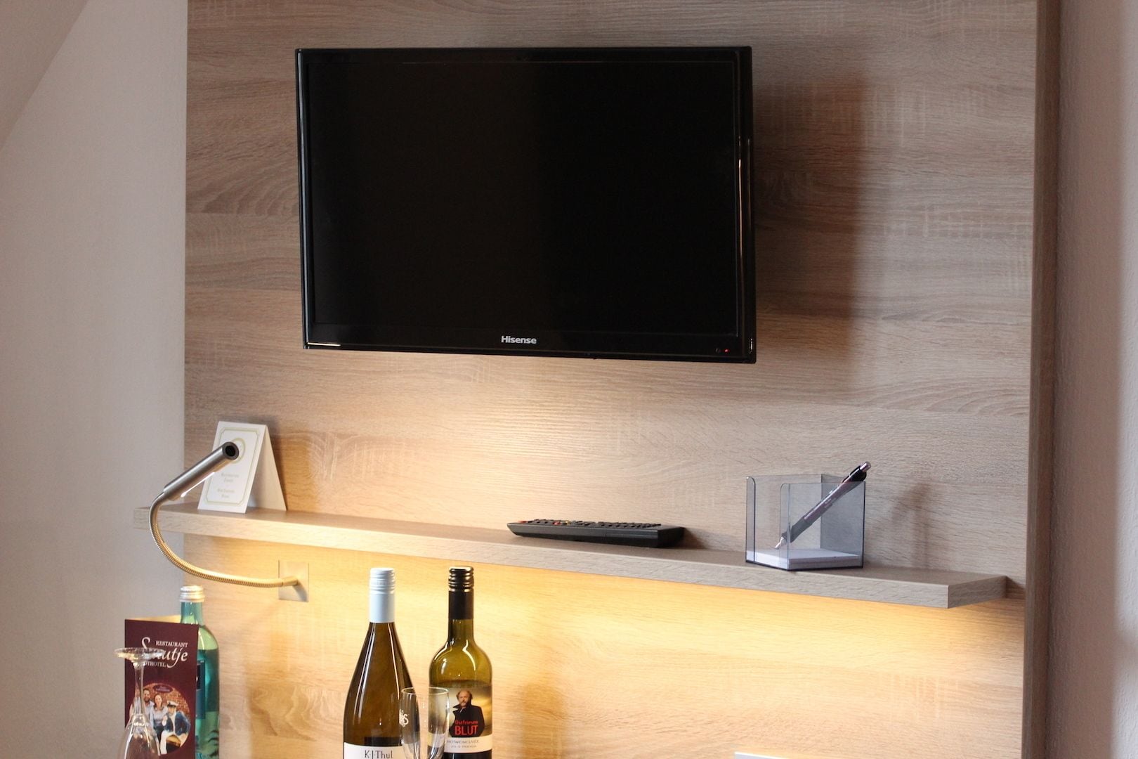Une chambre d’hôtel moderne équipée d’un téléviseur à écran plat fixé au mur, d’une étagère en bois sur laquelle se trouvent une télécommande, une bouteille de vin et un verre.