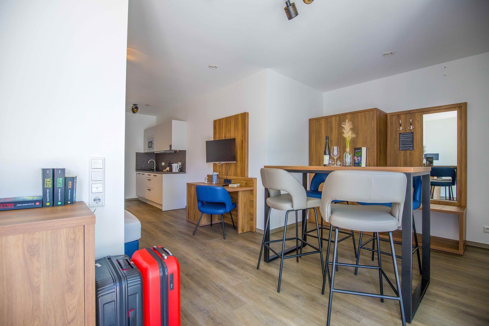 A compact hotel room with a kitchenette, a dining area with bar stools, a TV mounted on the wall, and a small desk.