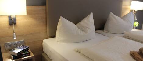 A neatly made bed with white linens and pillows, a bedside table with a lamp and a box, and a wall socket.