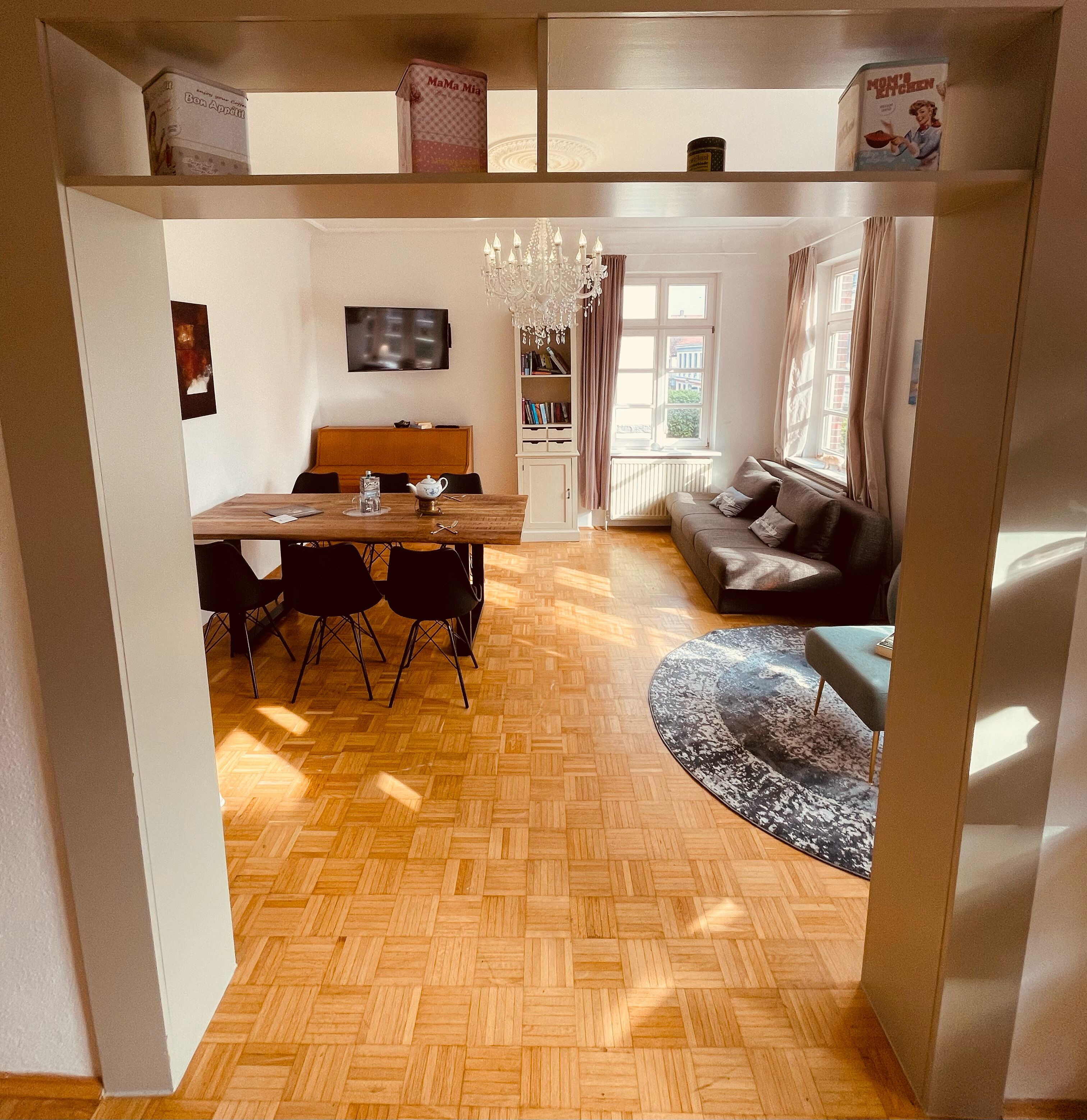 A dining area with a wooden table and chairs, a chandelier, and a bookshelf.