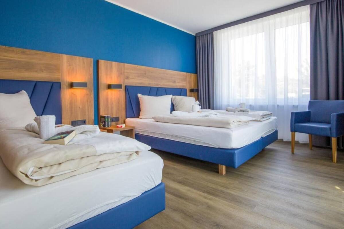 A hotel room with two beds, a blue accent wall, and a large window with curtains.