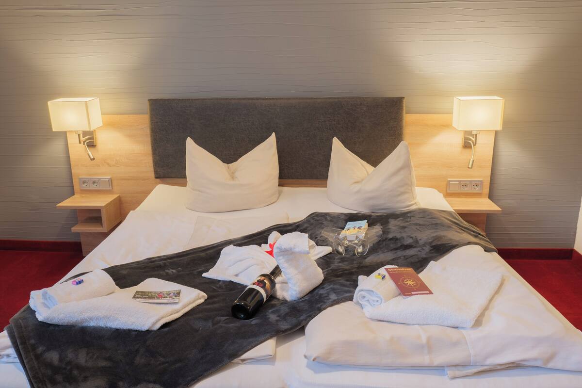 A neatly made bed with pillows, a blanket, towels, and a bottle of water. There are two bedside lamps and a wooden nightstand with a lamp.