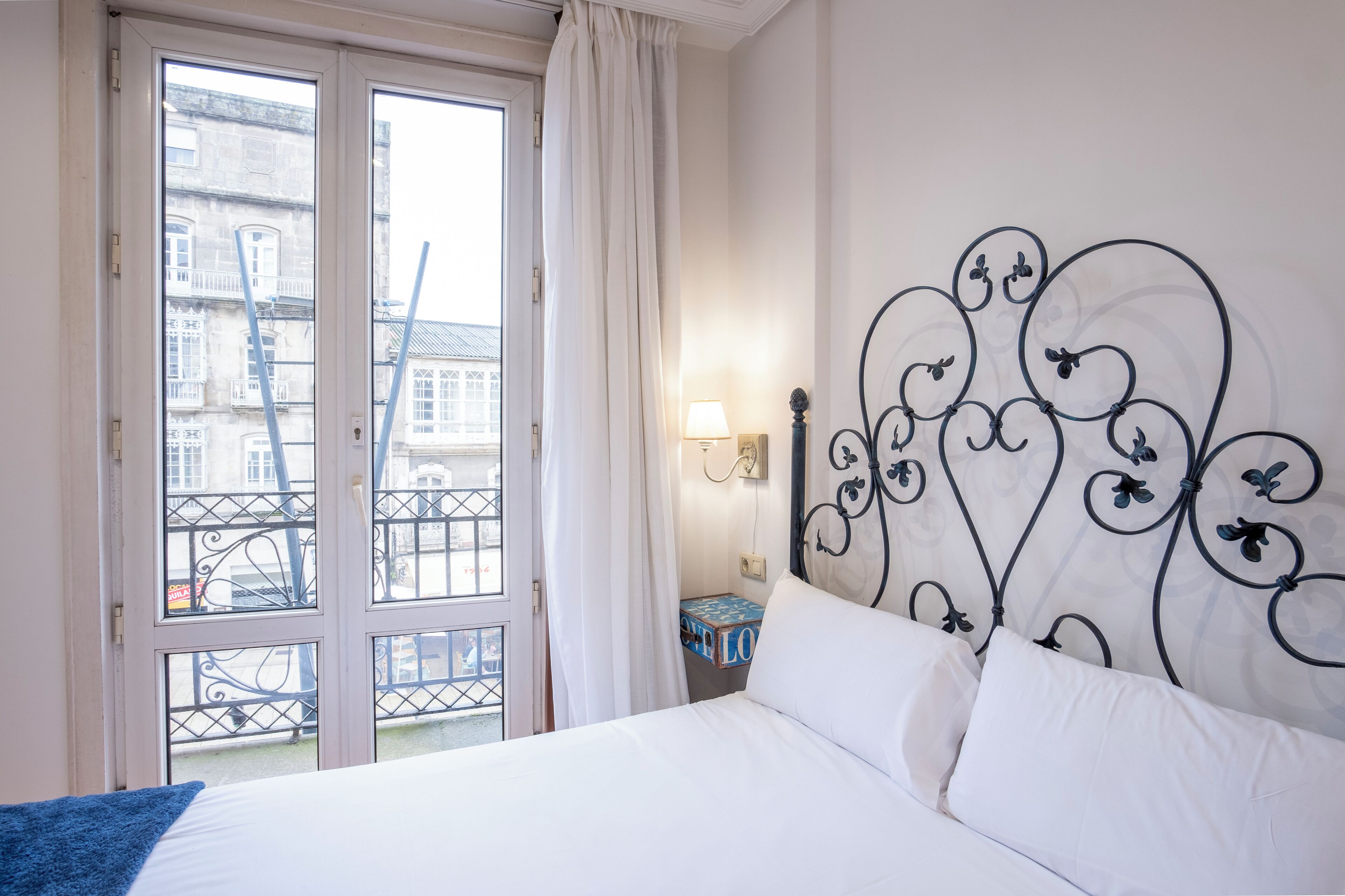 A hotel room with a bed, a window with curtains, a nightstand with a lamp, and a decorative metal headboard.