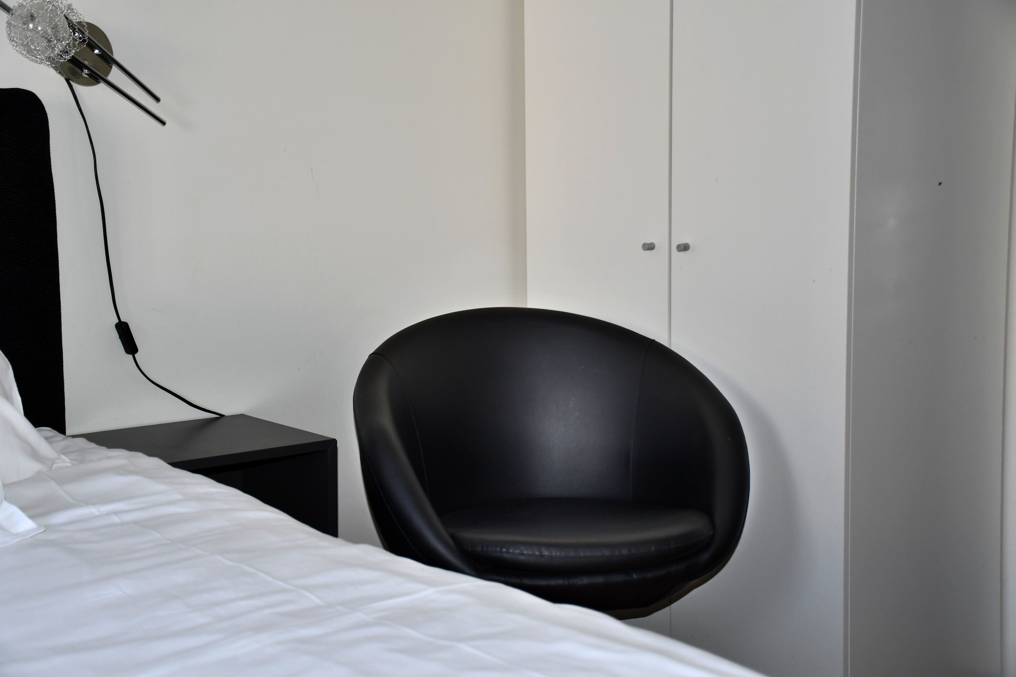 A modern hotel room with a bed, a black chair, a small table, and a wardrobe.