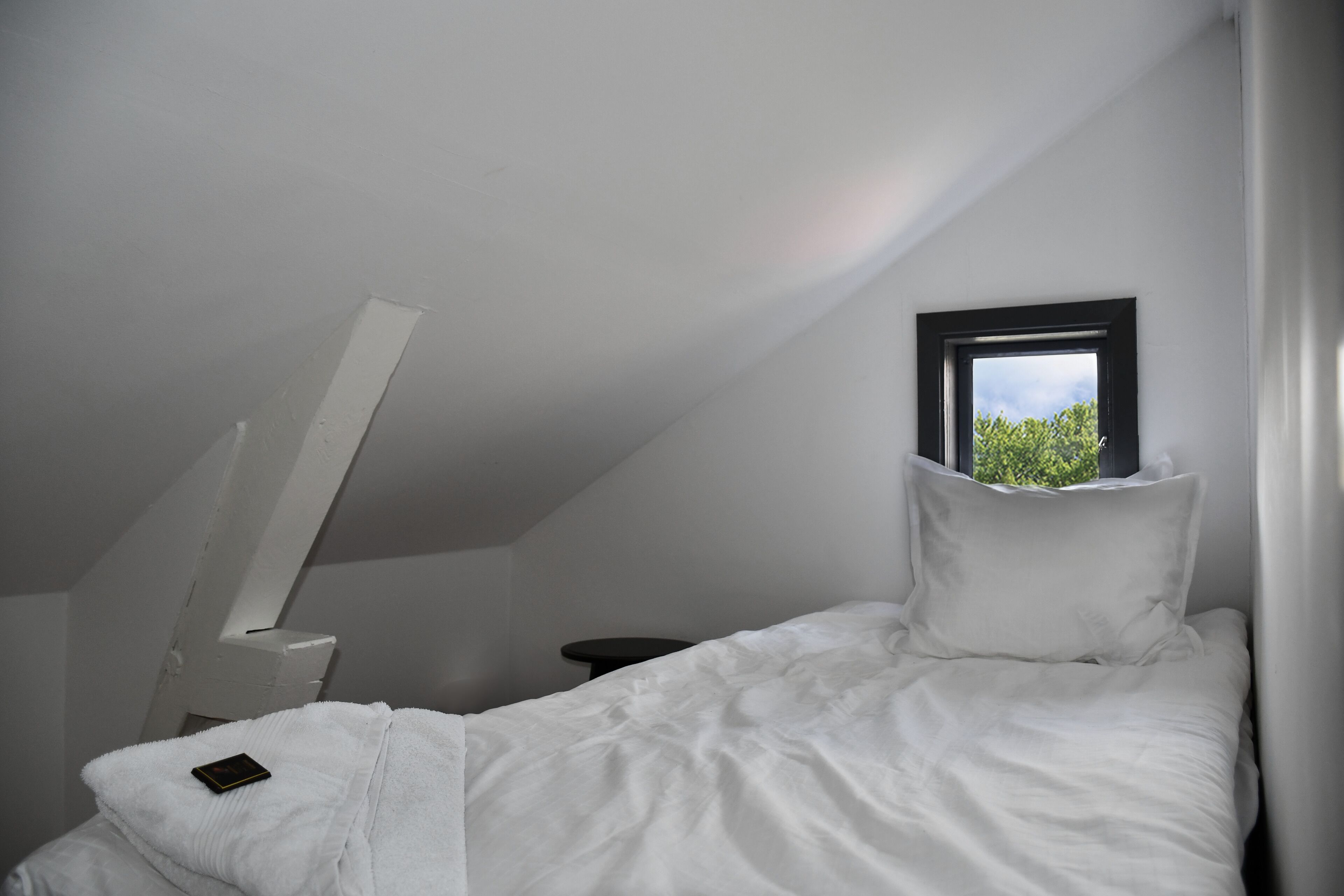 A bedroom with a sloped ceiling, a window, a bed with white bedding, and a small bedside table.