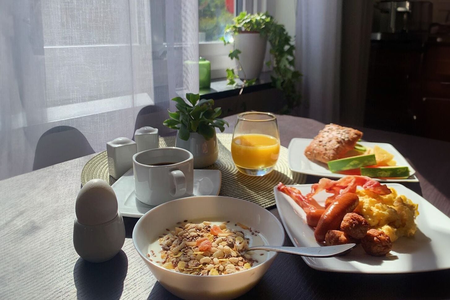 A breakfast setting with a bowl of cereal, a plate of eggs, toast, and juice on a table by a window.