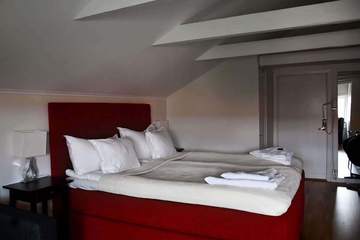 Hotellihuone, jossa on punainen sänky, valkoiset vuodevaatteet, yöpöytä lampun kanssa ja ovi oikealla puolella.