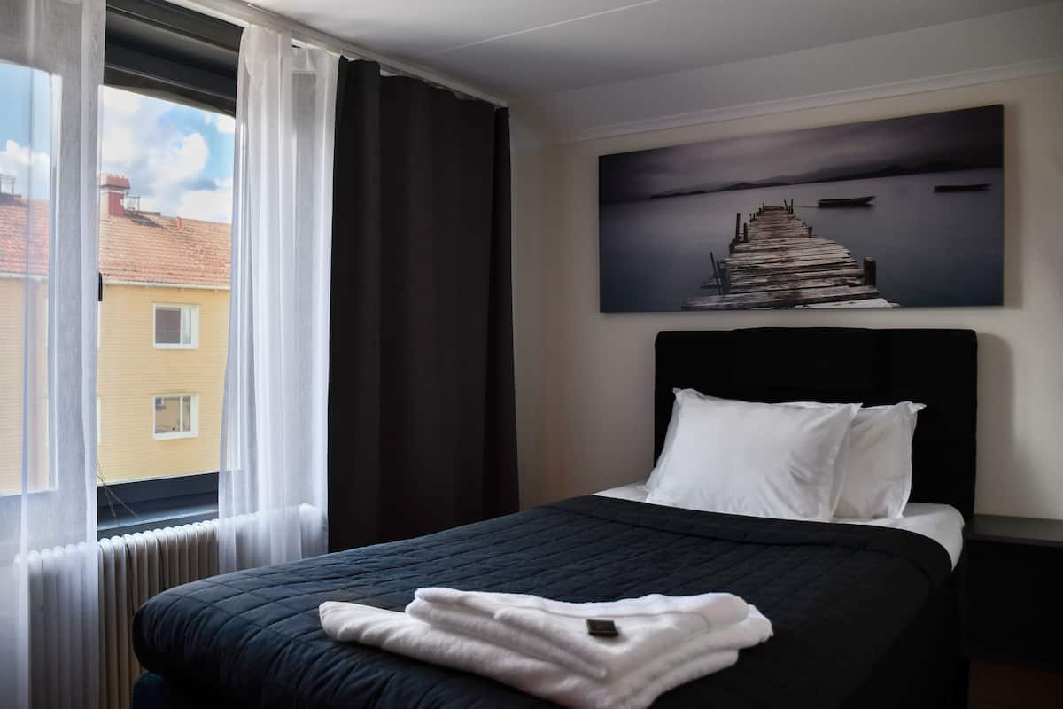 Hotellihuone, jossa on sänky, ikkunasta avautuva näkymä ympäröiviin rakennuksiin ja seinällä oleva taulu.