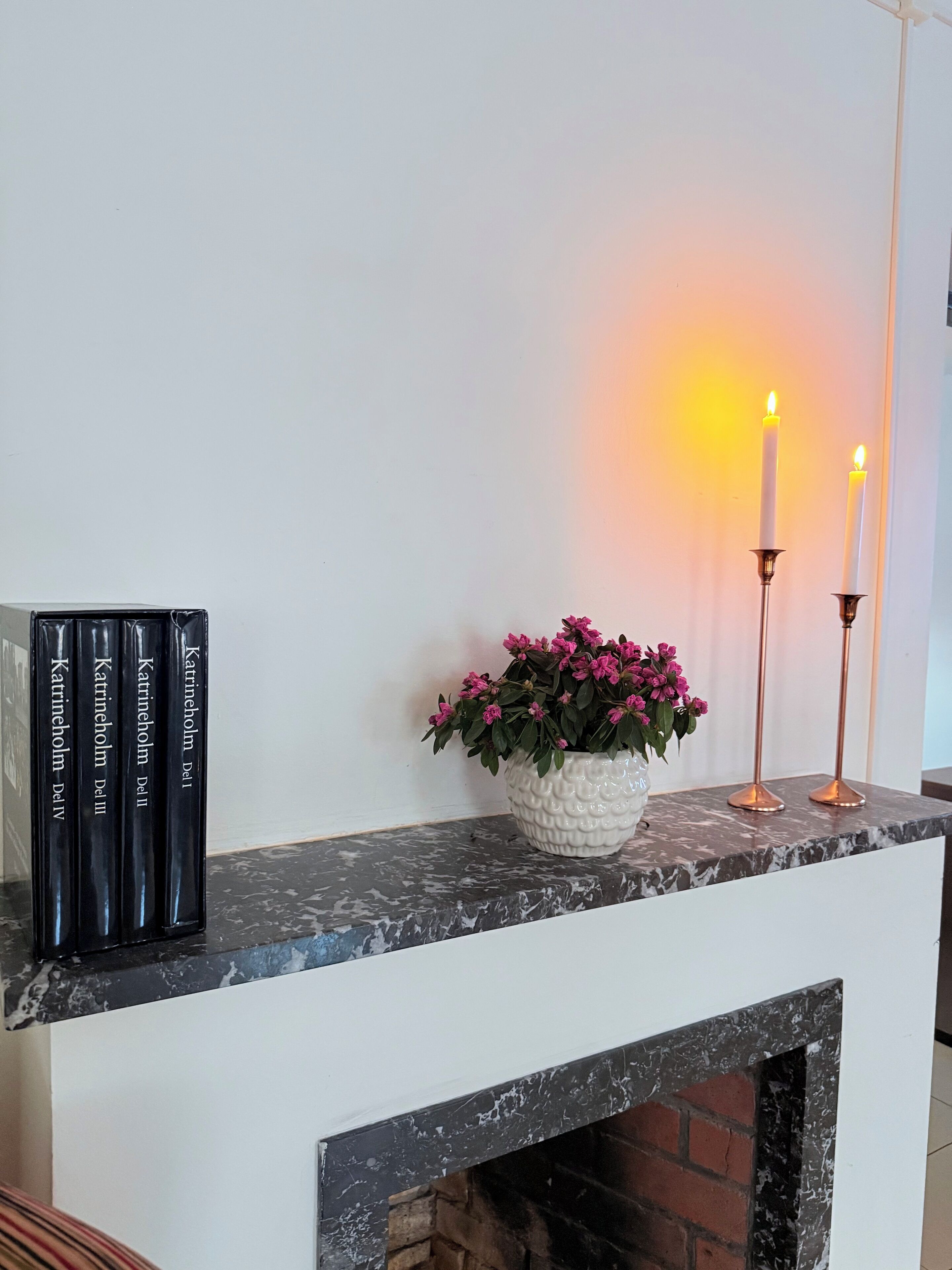 A fireplace mantel with a white vase of pink flowers, two lit candles, and a row of books titled 'Katrineholm Del II'.