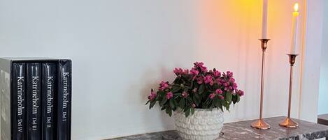 A fireplace mantel with a white vase of pink flowers, two lit candles, and a row of books titled 'Katrineholm Del II'.