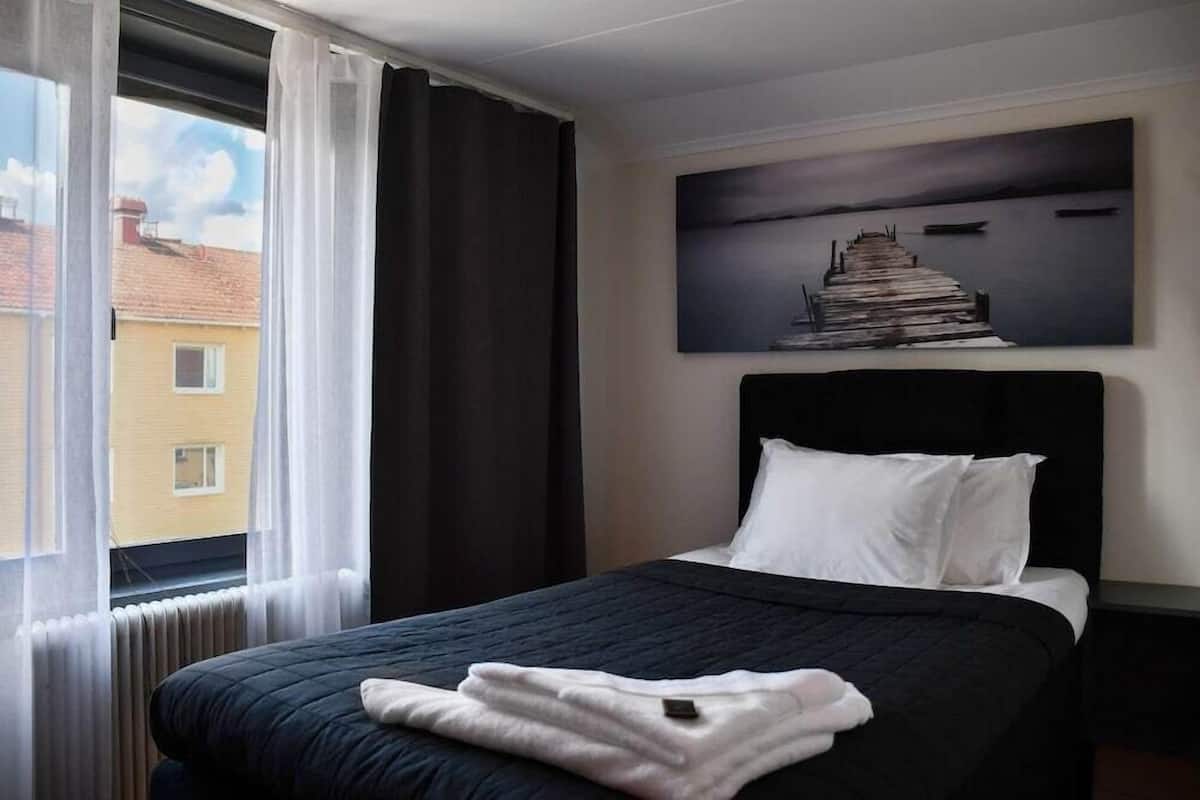 Hotellihuone, jossa on sänky, ikkuna verhoilla, näkymä rakennuksiin ja taulu seinällä.
