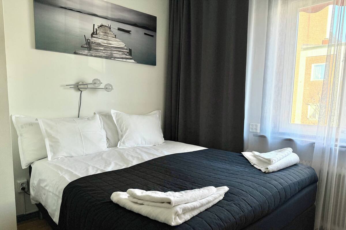 Hotellihuone, jossa on suuri sänky, valkoiset tyynyt, tumma, kuviollinen peitto ja seinällä kuva laiturista.