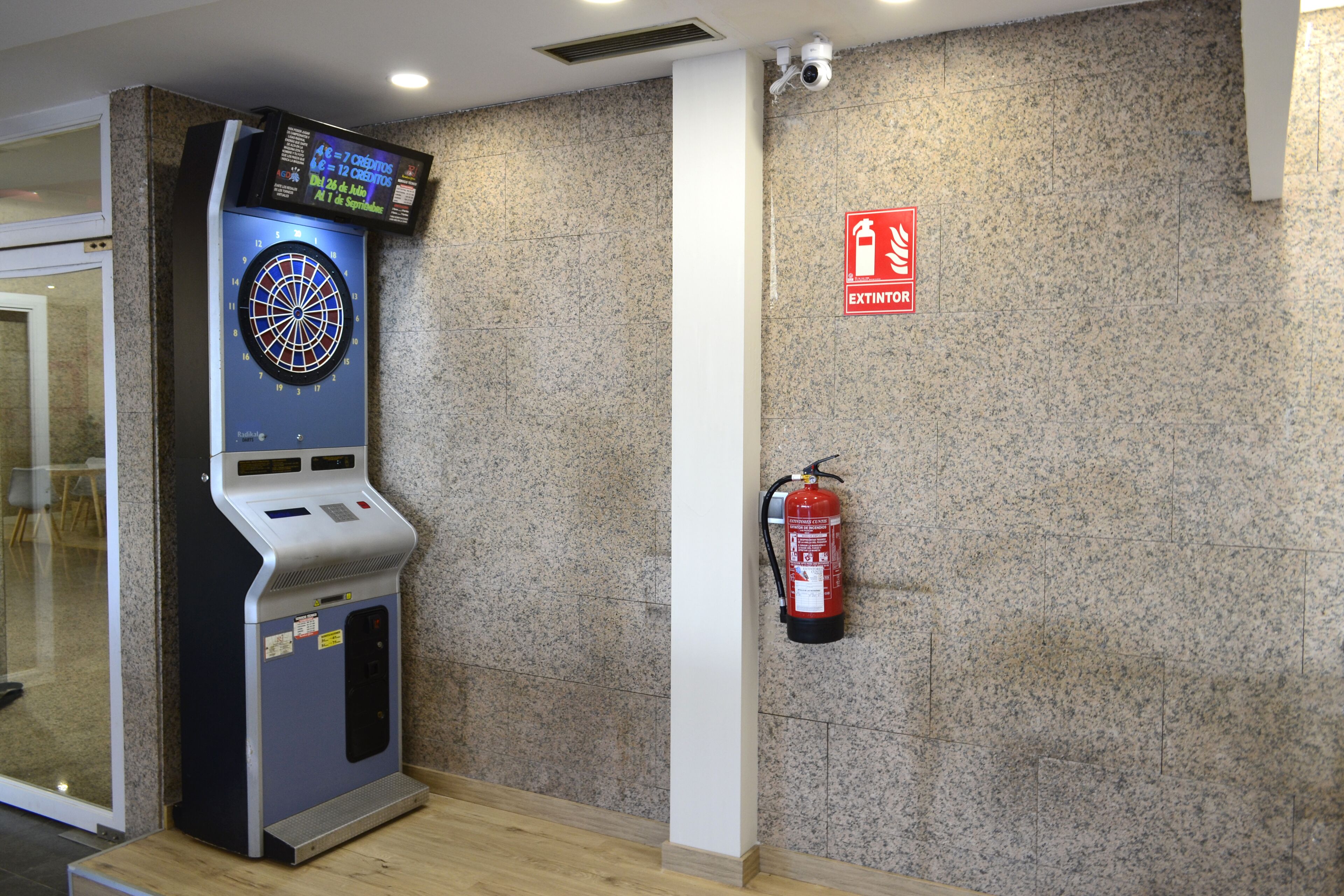 A dartboard, a fire extinguisher, and a digital display in a room with a tiled wall and a wooden floor.