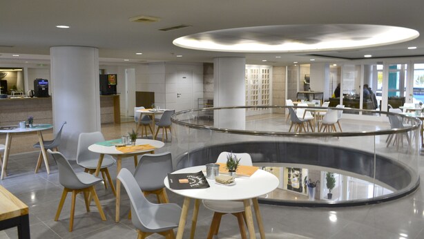 Un comedor moderno en el interior con mesas y sillas redondas, un elemento central de vidrio y una cocina visible al fondo.