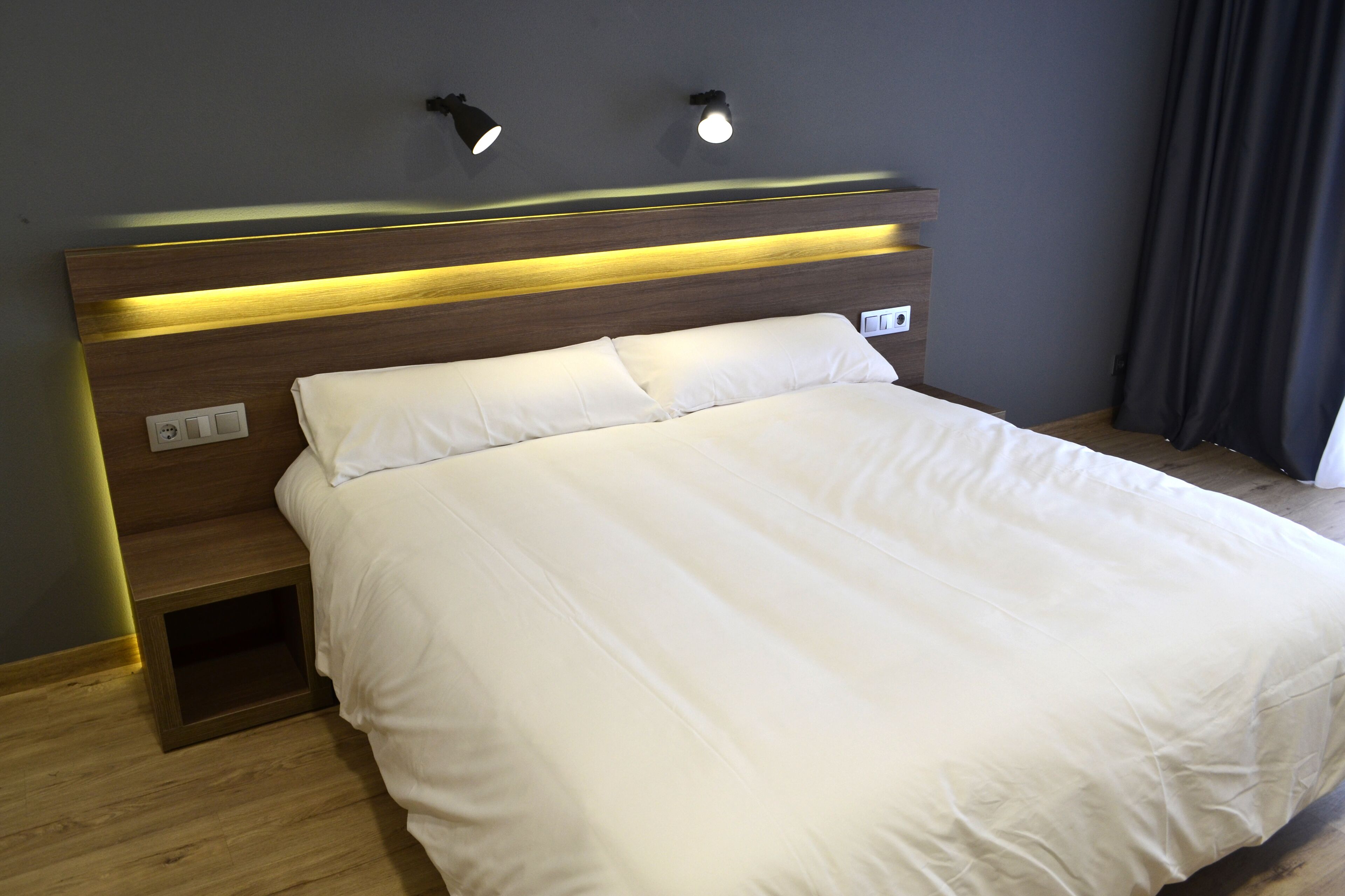 Un lit bien fait, avec du linge de lit blanc, dans une chambre moderne dotée d’une tête de lit en bois et d’un éclairage intégré.