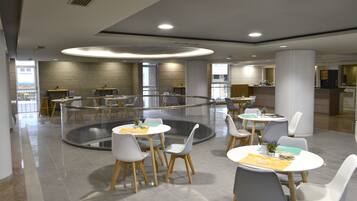 Un espace de restauration moderne avec des tables et des chaises rondes, une cloison en verre au centre et une vue sur la cuisine en arrière-plan.