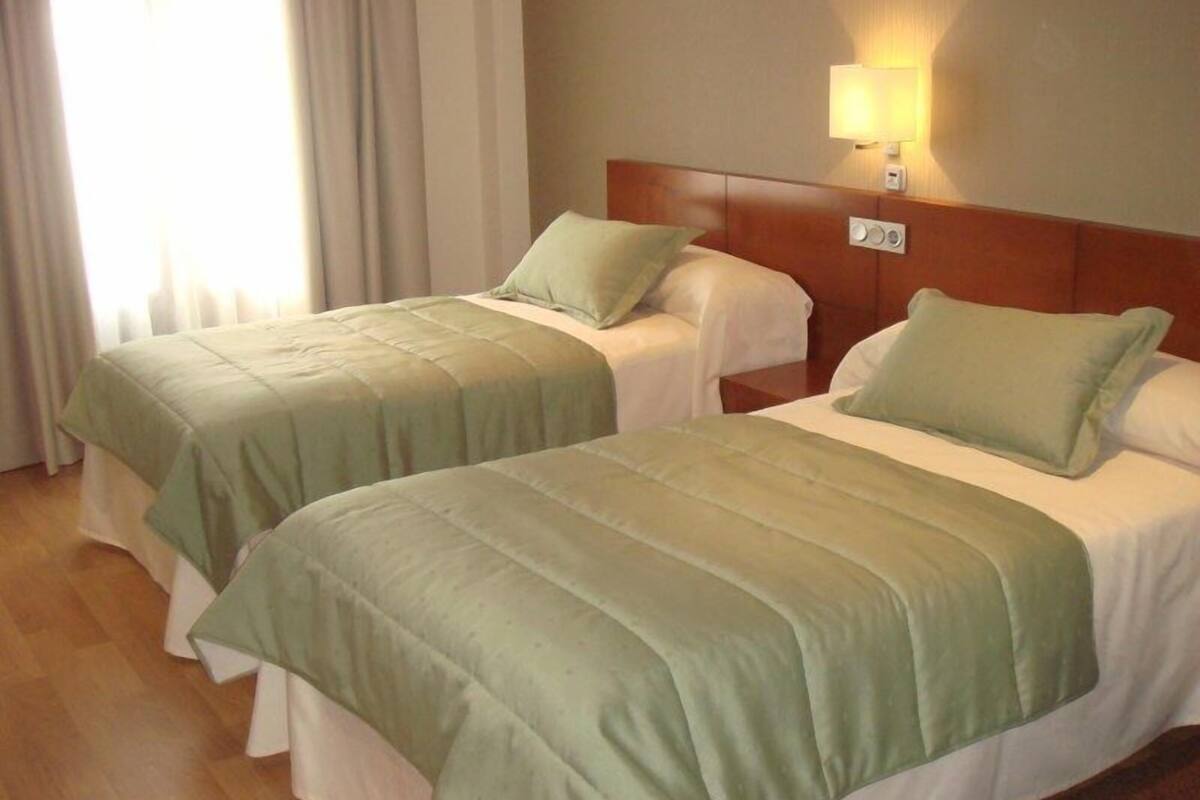 Two single beds with green bedding, a wooden headboard, a bedside lamp, and a window with curtains.