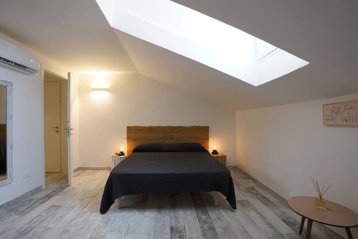A bedroom with a skylight, a bed with a dark headboard, bedside lamps, and a small round table with a candle.