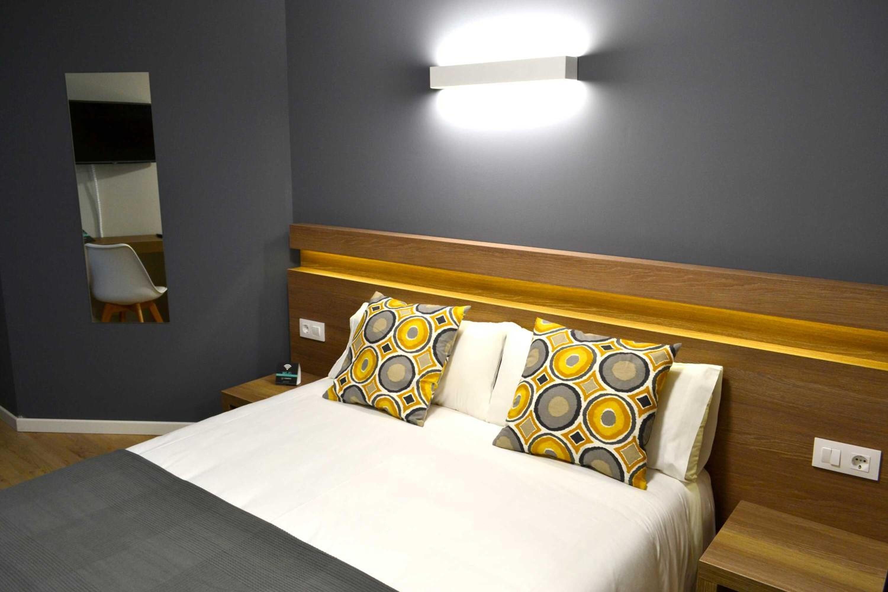 Una cama bien hecha con almohadas estampadas, una mesita de noche y una lámpara fijada a la pared.