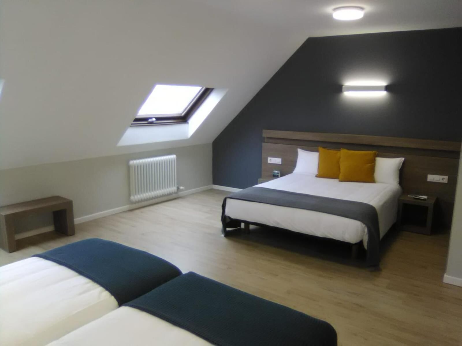 Een slaapkamer met een schuine plafond, dakraam, bed met wit en geel beddengoed, nachtkastje en een nachttafel.