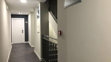 A narrow hallway with a carpeted floor, a white door, a staircase with a metal railing, and a fire alarm on the wall.