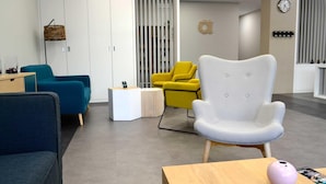 Een moderne wachtruimte met een blauwe bank, een gele fauteuil, een witte fauteuil, een kleine ronde tafel en een houten kast.