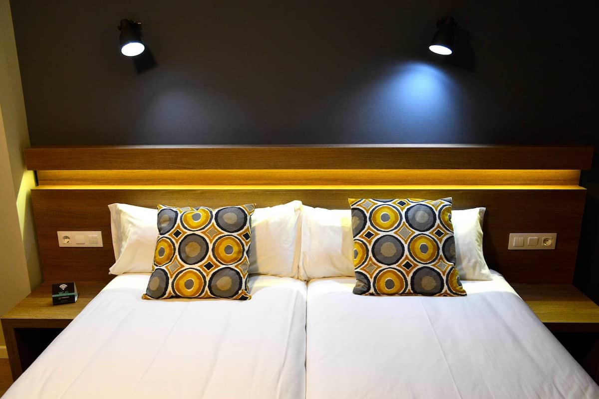 Un lit double avec une literie blanche et des coussins à motifs, une tête de lit en bois et deux lampes fixées au mur.