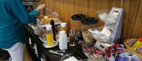 Una barra de desayuno con diversos alimentos, una máquina de café y un televisor mostrando un evento deportivo.