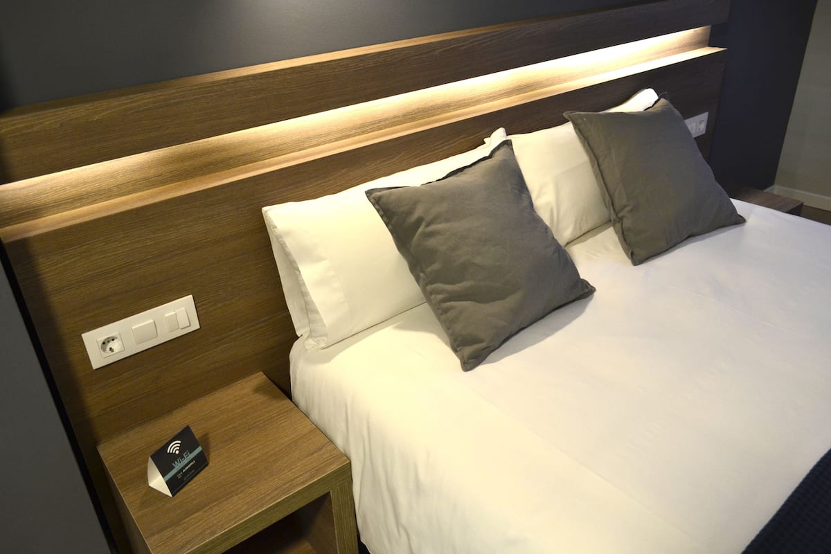 Un lit bien fait, avec du linge de lit blanc et gris, une tête de lit en bois et une table de chevet sur laquelle se trouve un petit appareil électronique.