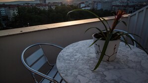 Een balkon met een bloempot en een metalen stoel, met uitzicht op een stadslandschap bij zonsondergang.