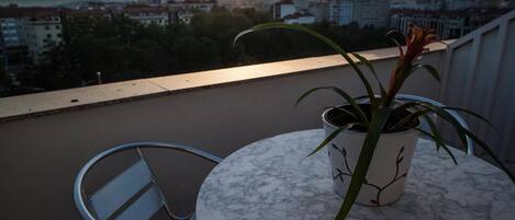 Un balcón con una planta en maceta y una silla metálica, con vistas a un paisaje urbano al atardecer.