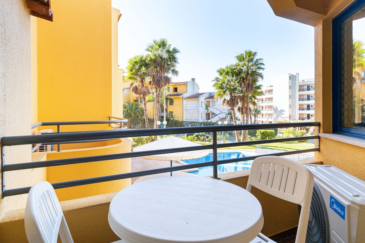 Varanda com mesa e cadeiras, vista para uma piscina e palmeiras.