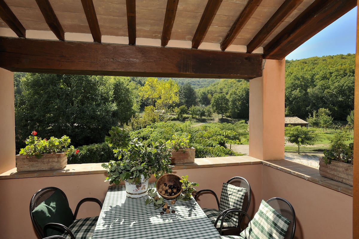 A covered outdoor seating area with a table and chairs, surrounded by potted plants and a scenic view of a vineyard.