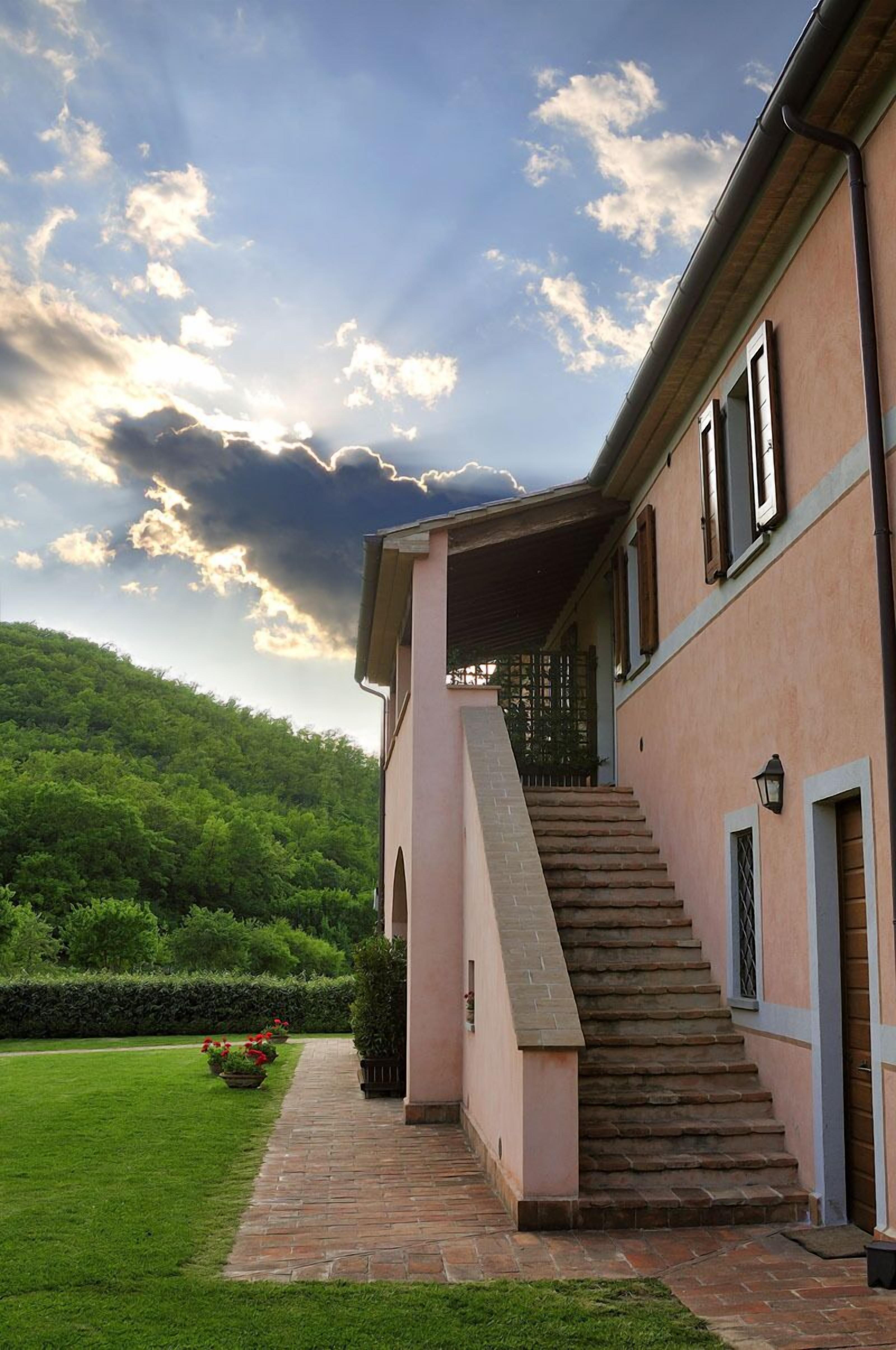A building with a staircase leading to a covered porch, surrounded by a well-maintained lawn and a hill in the background.