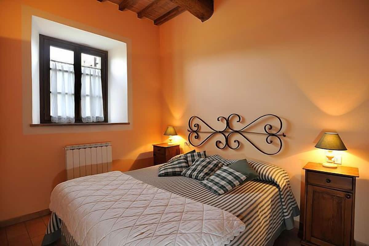 A bedroom with a bed, two bedside tables with lamps, a radiator, and a window with curtains.
