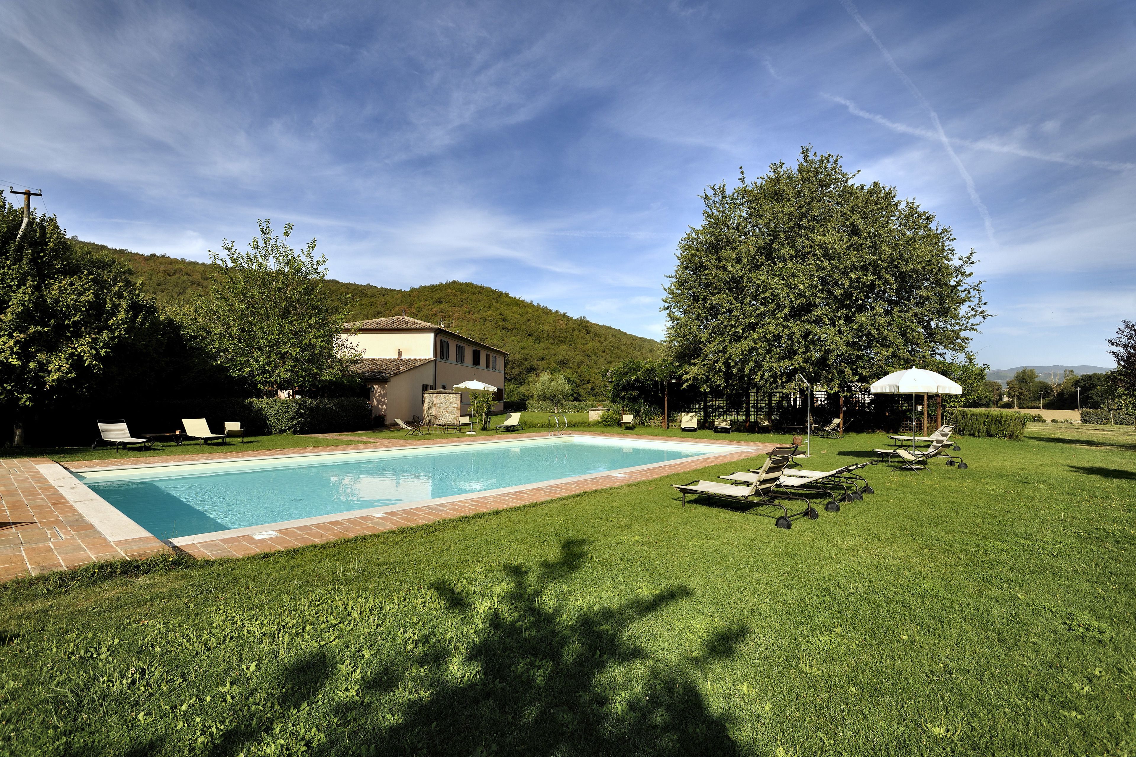 A swimming pool with lounge chairs and umbrellas, surrounded by a well-maintained lawn and a hill in the background.