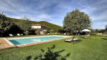 A swimming pool with lounge chairs and umbrellas, surrounded by a well-maintained lawn and a hill in the background.