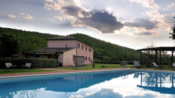 Une piscine entourée de chaises longues, un bâtiment doté d’un toit en tuiles et, au loin, une colline boisée.