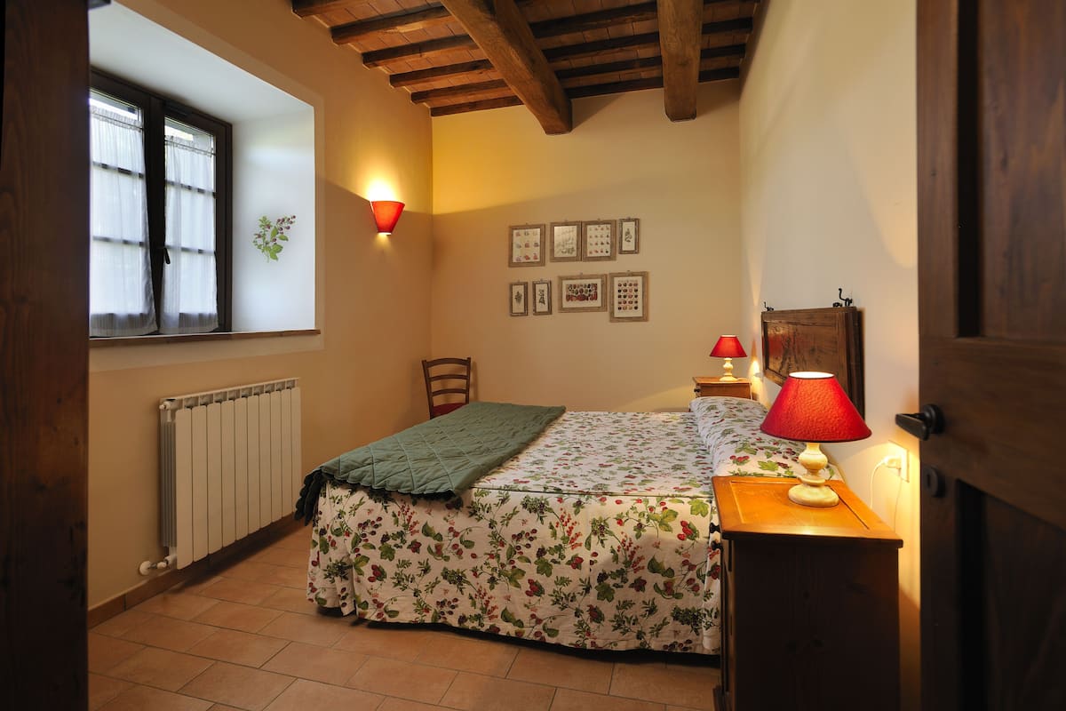 A bedroom with a bed, a nightstand, a lamp, a window with curtains, a radiator, and framed pictures on the wall.