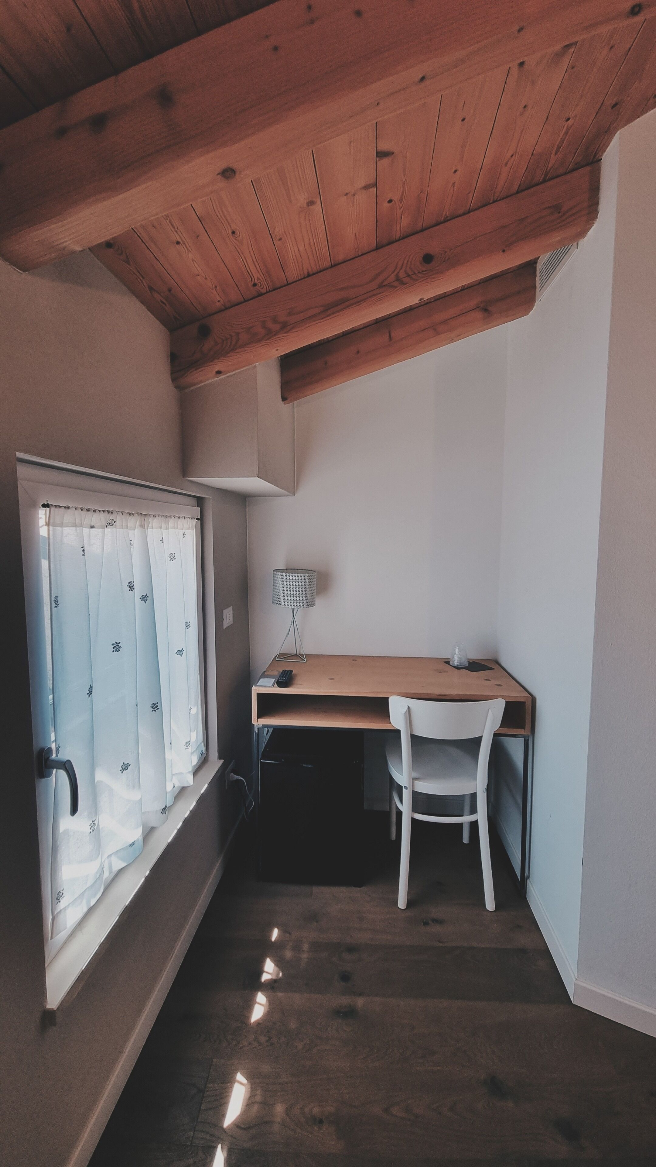 A small, compact room with a wooden ceiling, a desk with a chair, a window with curtains, and a lamp.