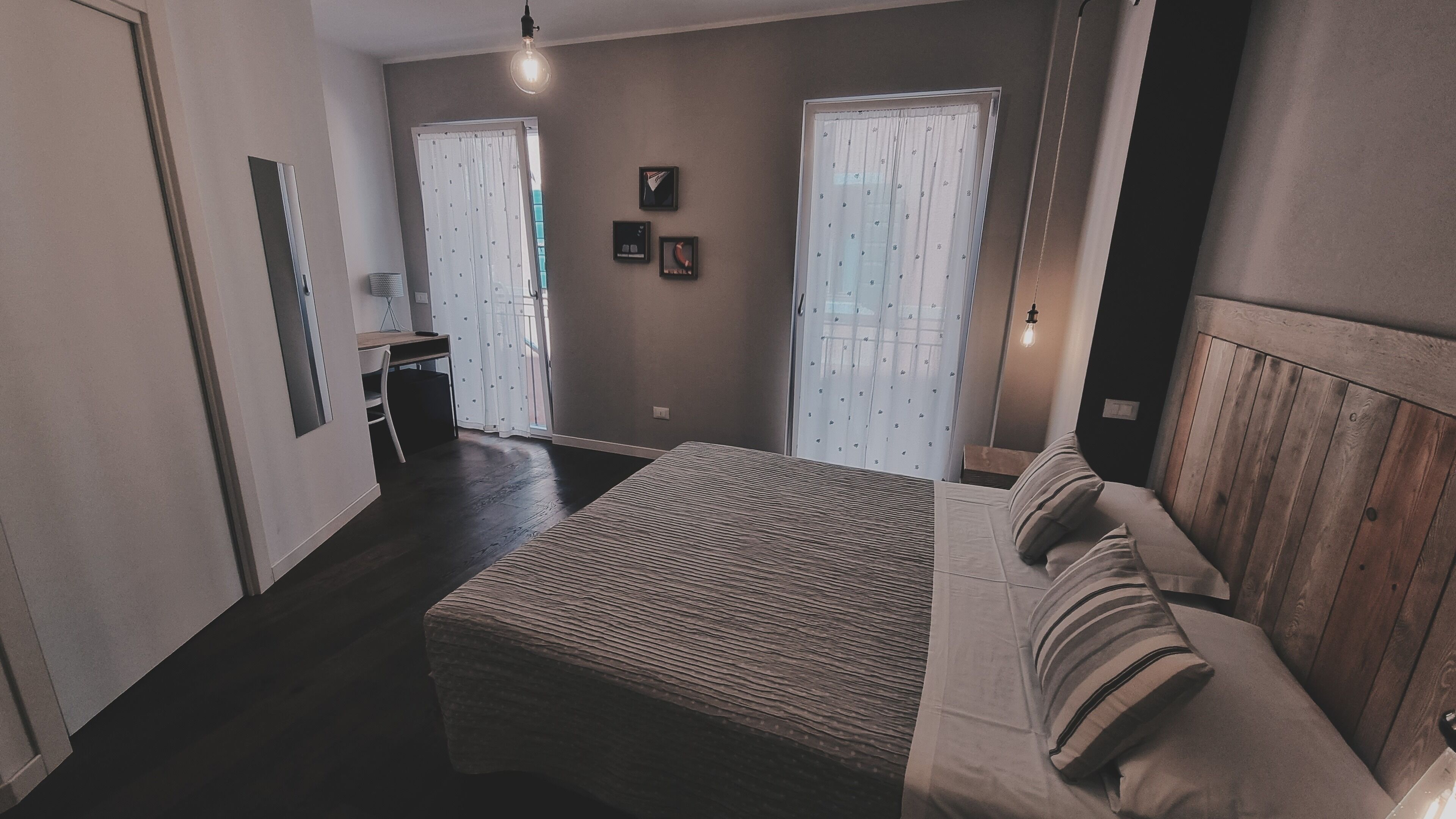 Een slaapkamer met een bed, een houten hoofdbord, een nachtkastje en een stoel. Er zijn twee ramen met gordijnen, een wandlamp en een klein plankje met decoratieve spullen.