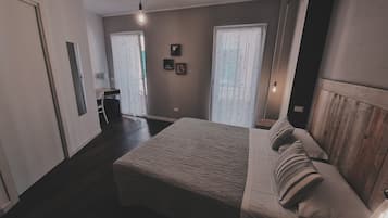 Een slaapkamer met een bed, een houten hoofdbord, een nachtkastje en een stoel. Er zijn twee ramen met gordijnen, een wandlamp en een klein plankje met decoratieve spullen.