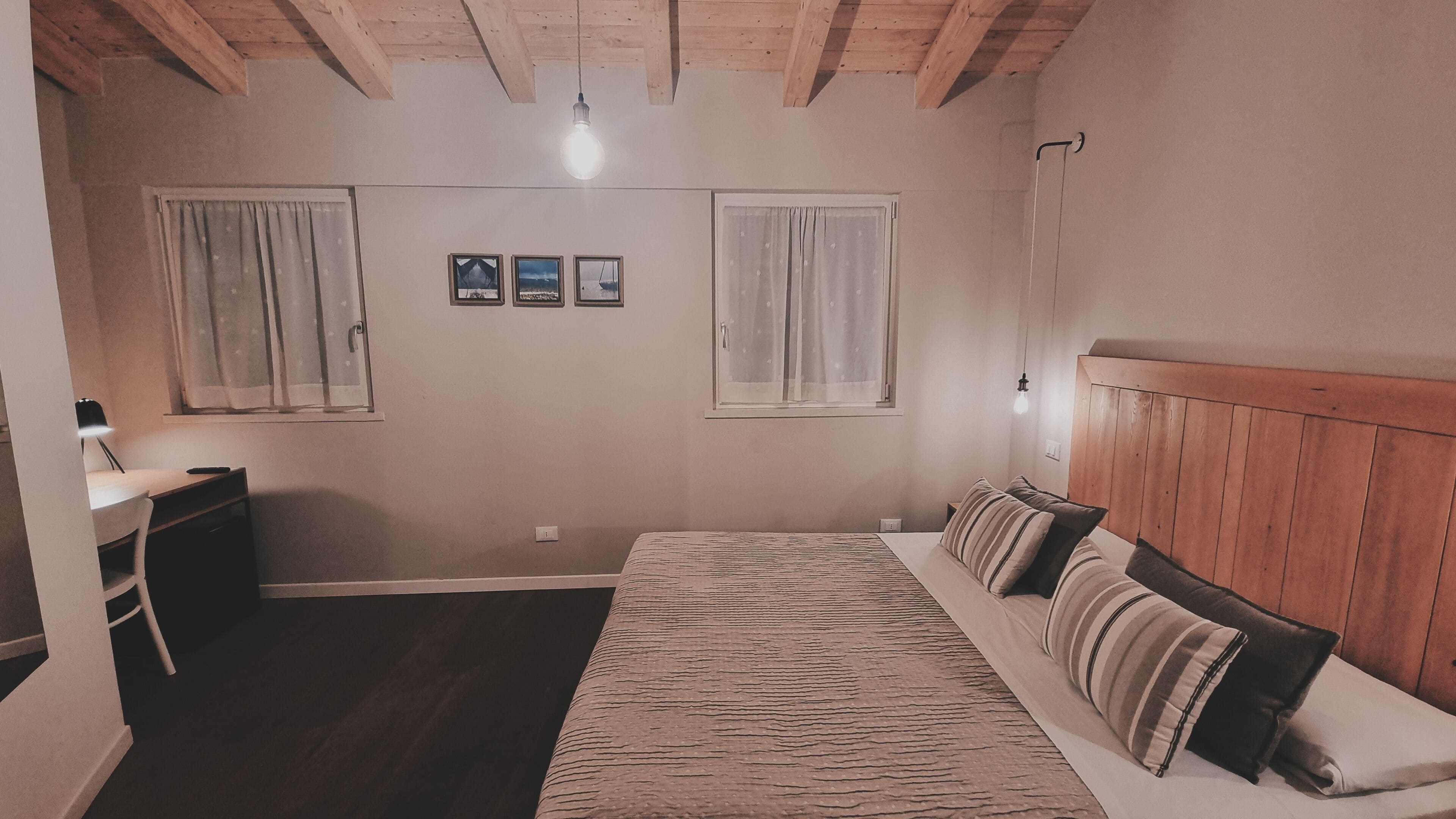 Una camera da letto con un letto, due finestre con tende, una scrivania con una lampada e un poggiatesta in legno.