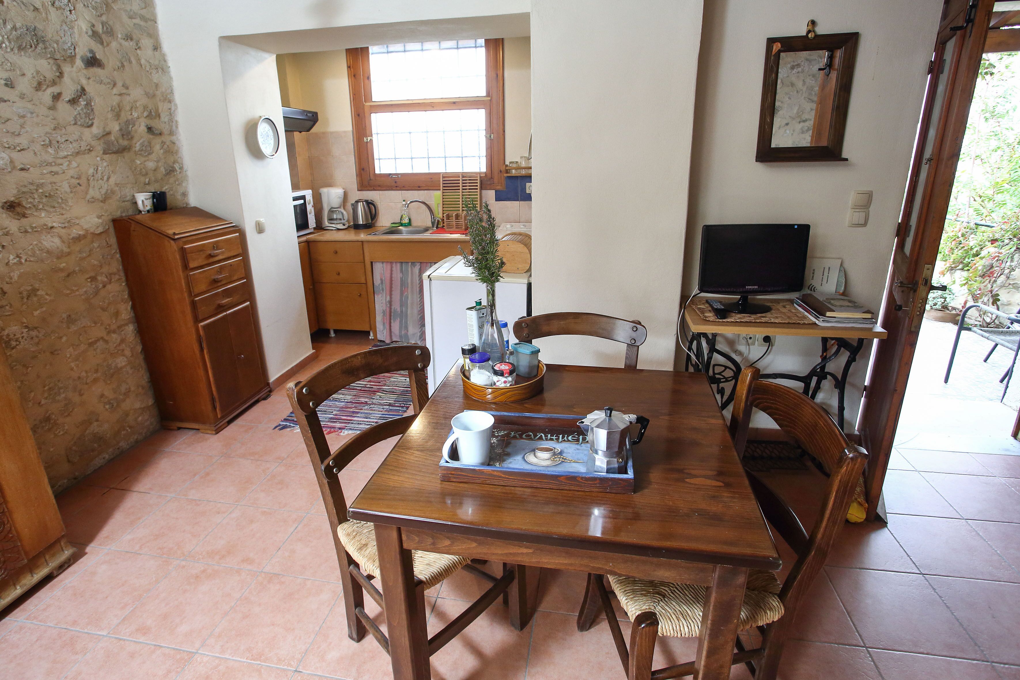 A dining area with a wooden table, chairs, a small kitchen in the background, and a window.