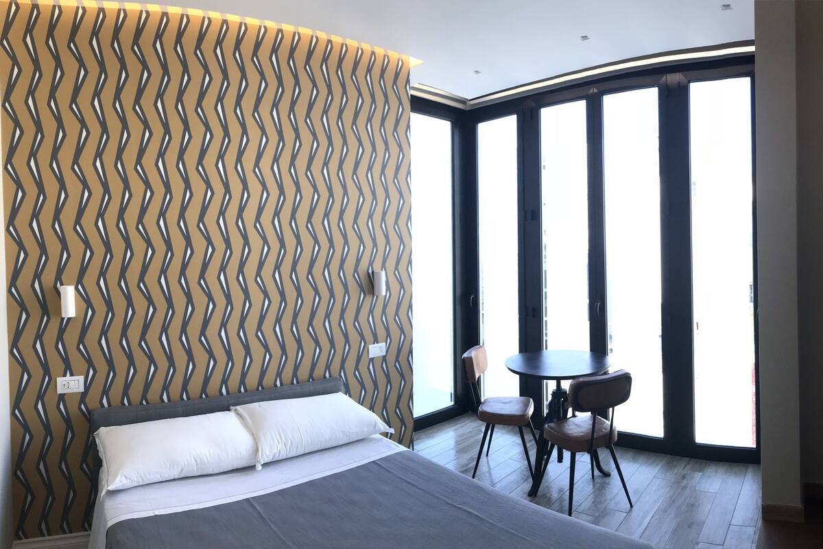 A hotel room with a bed, a small table with chairs, a TV, and large windows.
