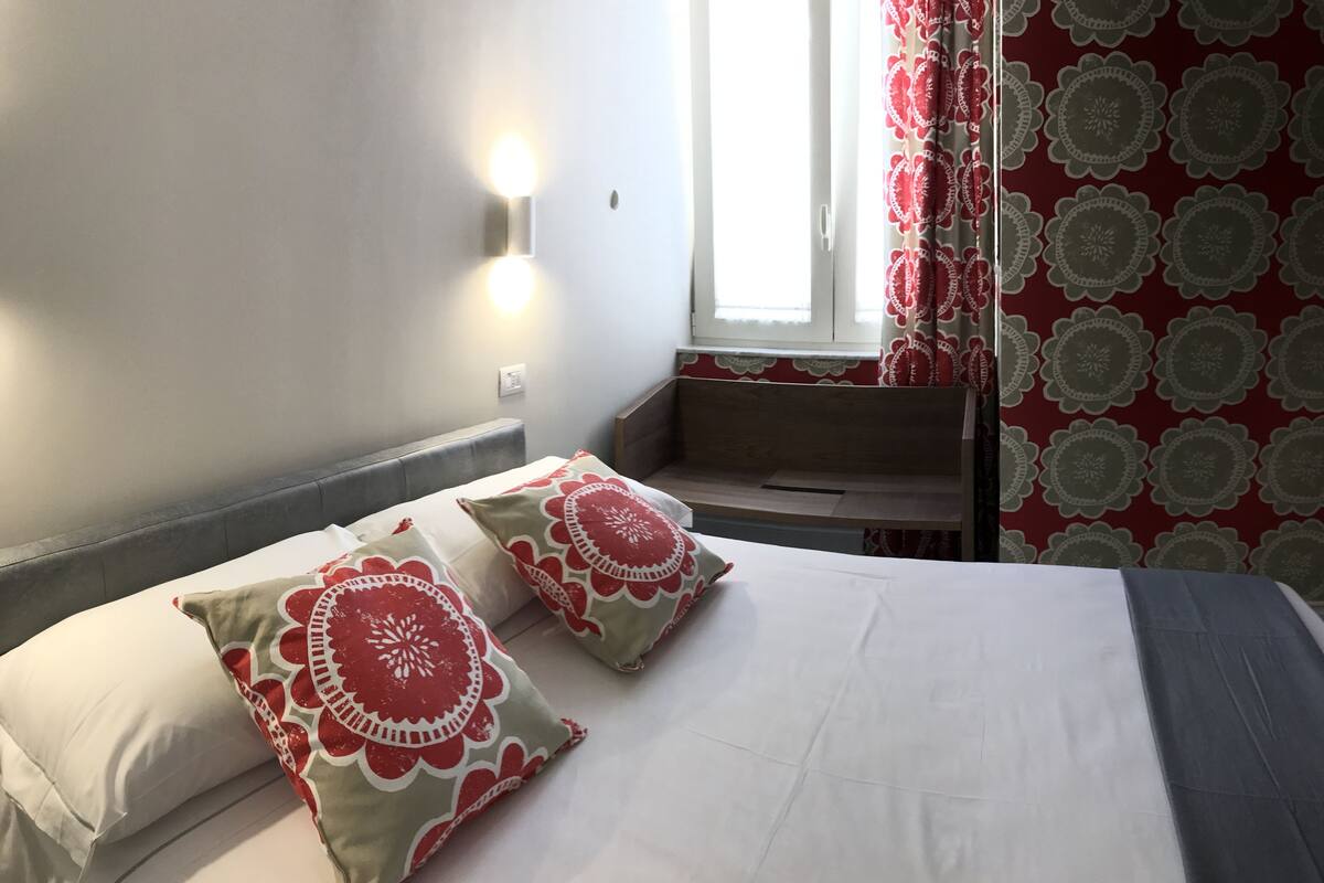 A hotel room with a bed, two pillows, a nightstand, a window with curtains, and a wall with a light fixture.