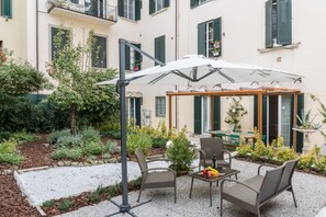An outdoor patio with wicker furniture, a white umbrella, and a garden area with various plants and a small tree. - Pilastri Garden by Mmega (Florence)