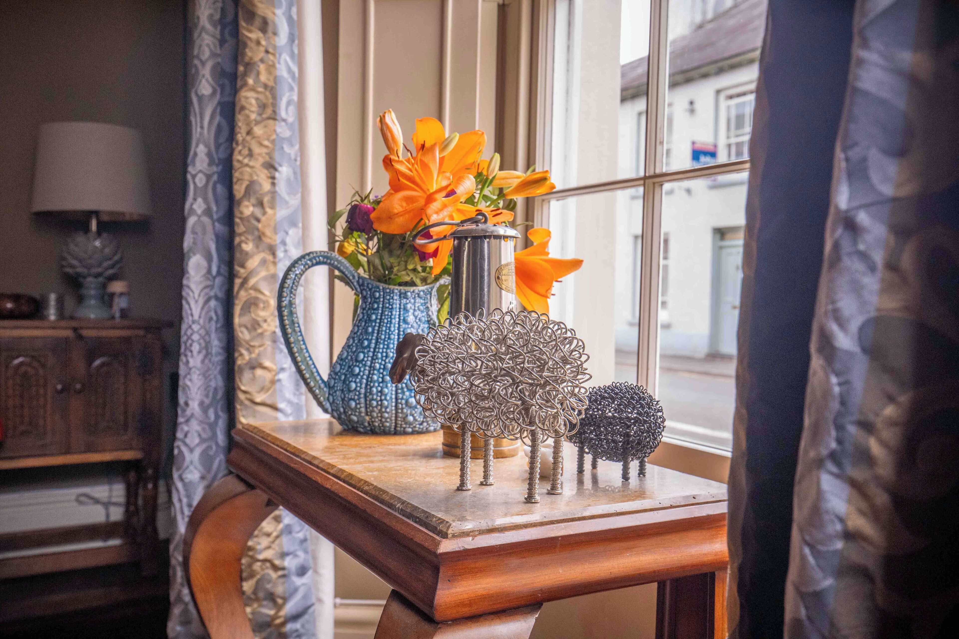 A wooden table with a blue pitcher, orange flowers, and metal tree sculptures.
