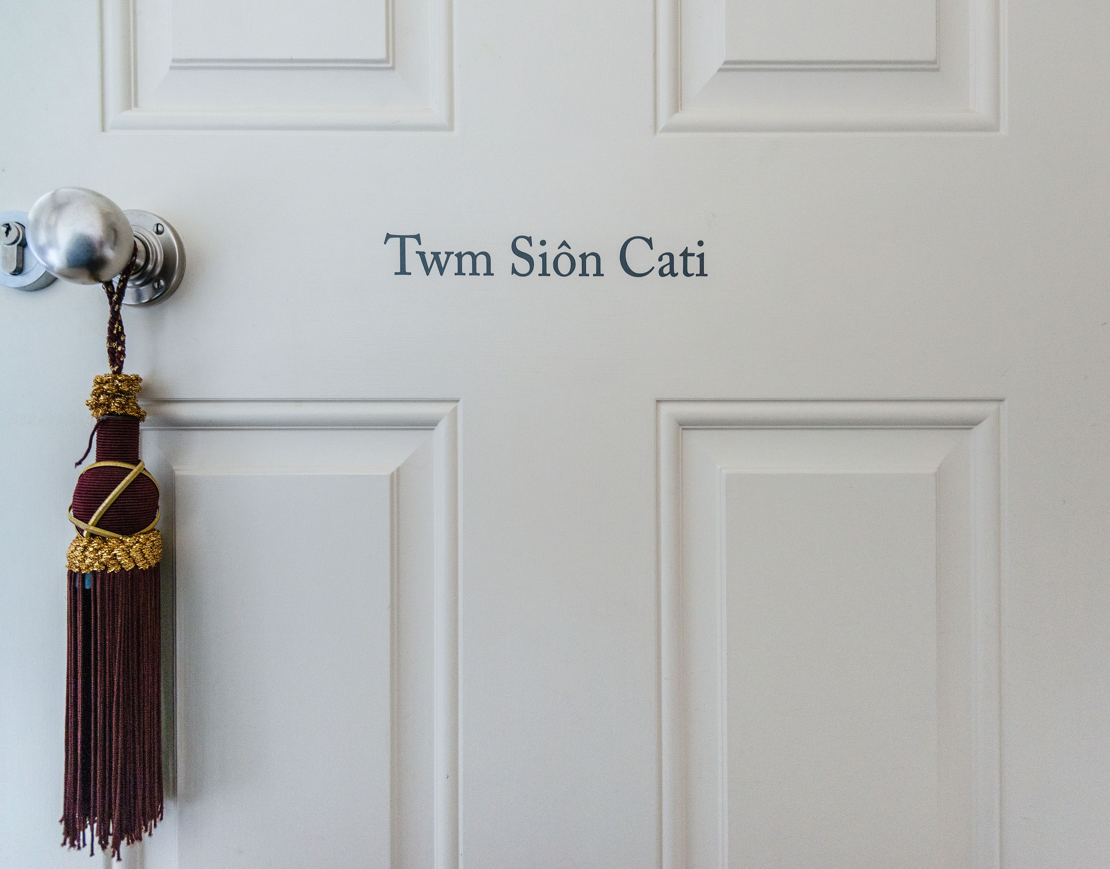 En hvid dør med en nøglehul og en dekorativ dusk. Teksten "Twm Siôn Cati" er synlig på døren.