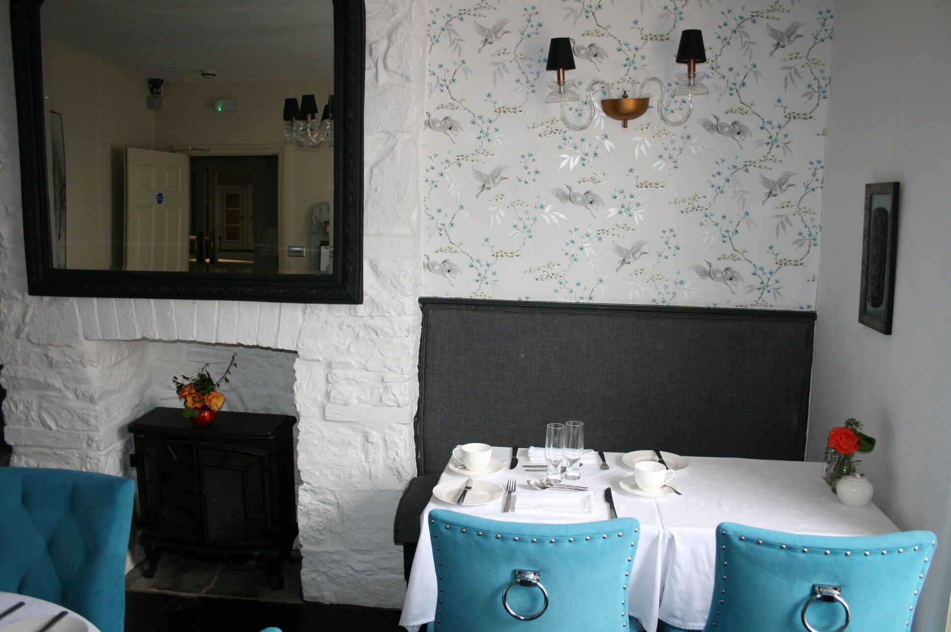 A dining area with a white table set for two, two blue chairs with metal rings, a white wall with floral wallpaper, a black-framed mirror, and a small vase with a red flower.